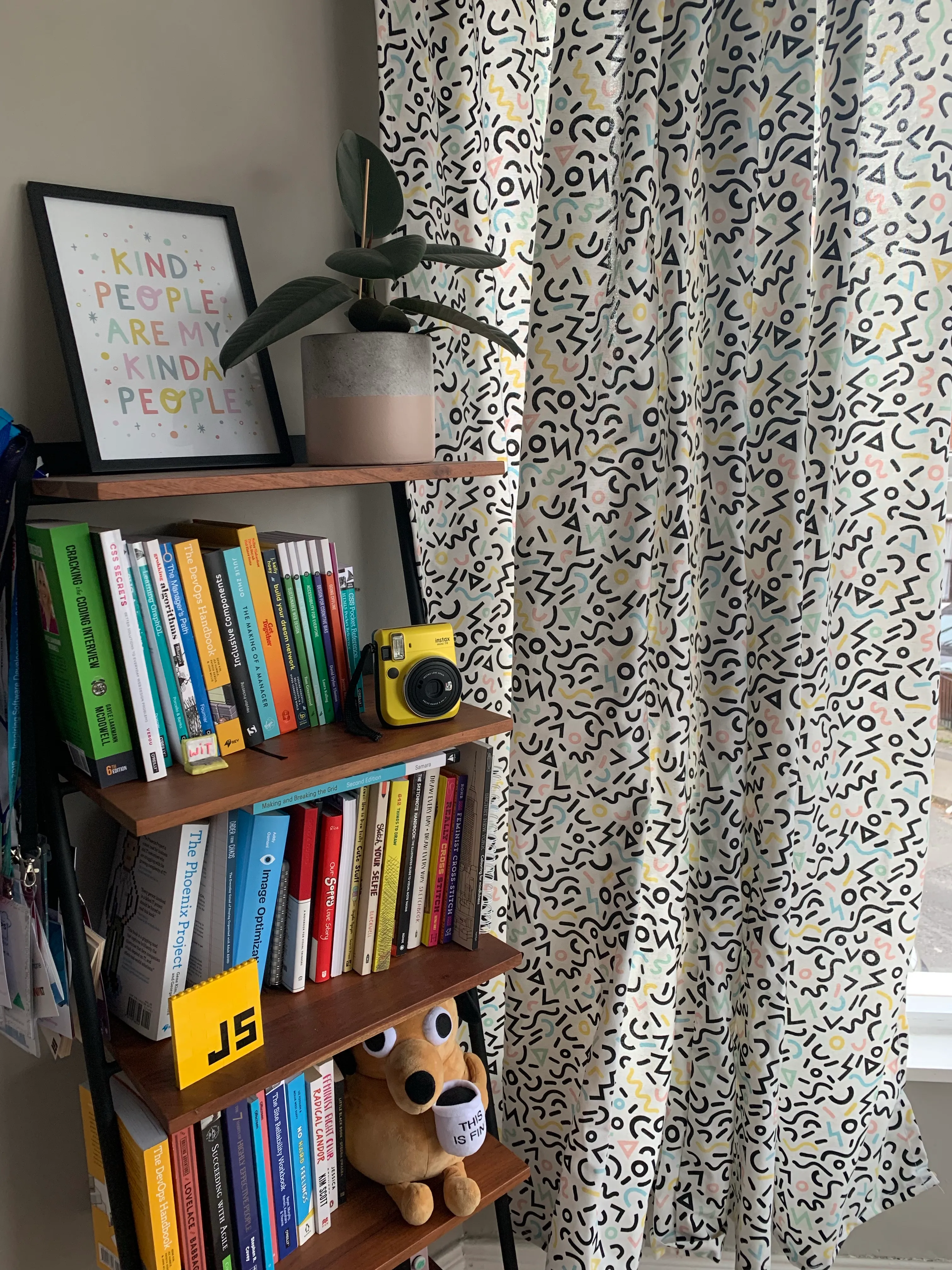 My bookshelf by the window in my office at home, you can see a bunch of tech focussed books and the curtain on the right hand side. The curtain is made of white canvas with a geometric print, that has mostly black shapes like triangles and circles and a small portion of the shapes are printed in pastel colours.