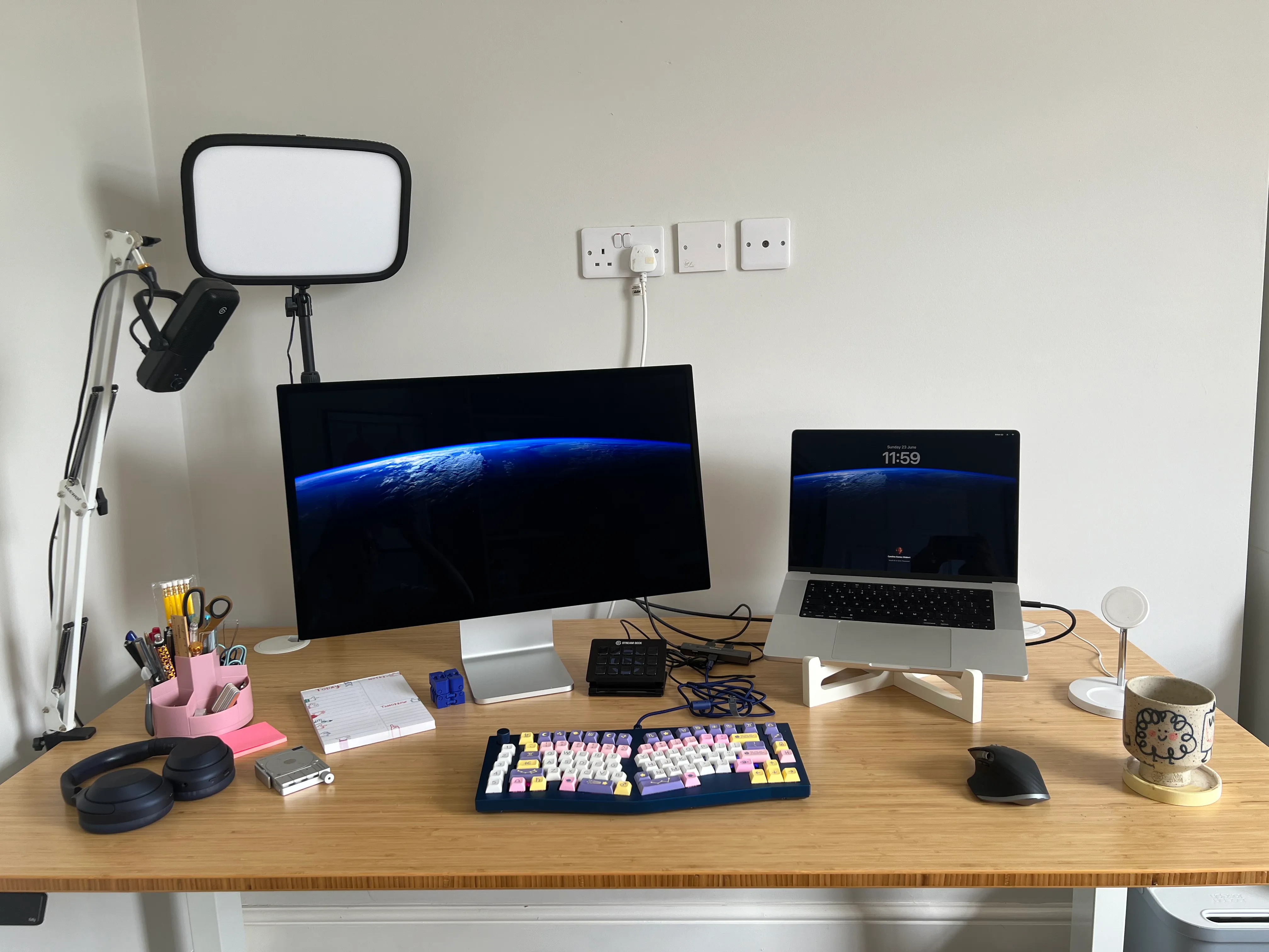 My desk, a bamboo top with a 27 inch monitor, and a laptop on a white laptop stand. You can also see on the desk a notepad, a pink pencil cup, a stream deck, a wireless phone charger, a coffee mug on top of a yellow coaster. Attached to the desk are a big rectangular keylight and a microphone on a boom arm. 