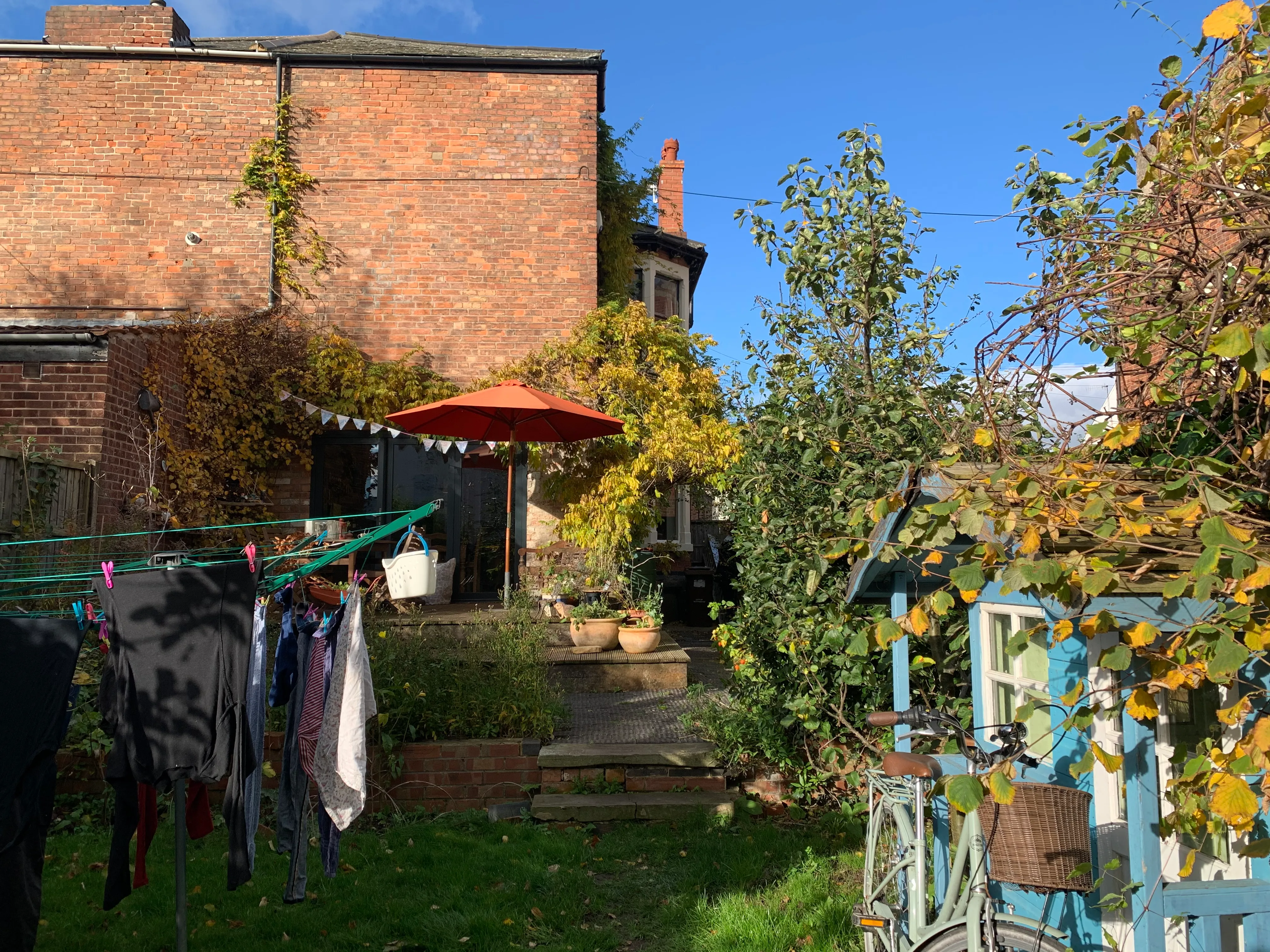 You can the back of a two storey brick house, a deck with a table and a parasol, some bunting and a blue wendy house.