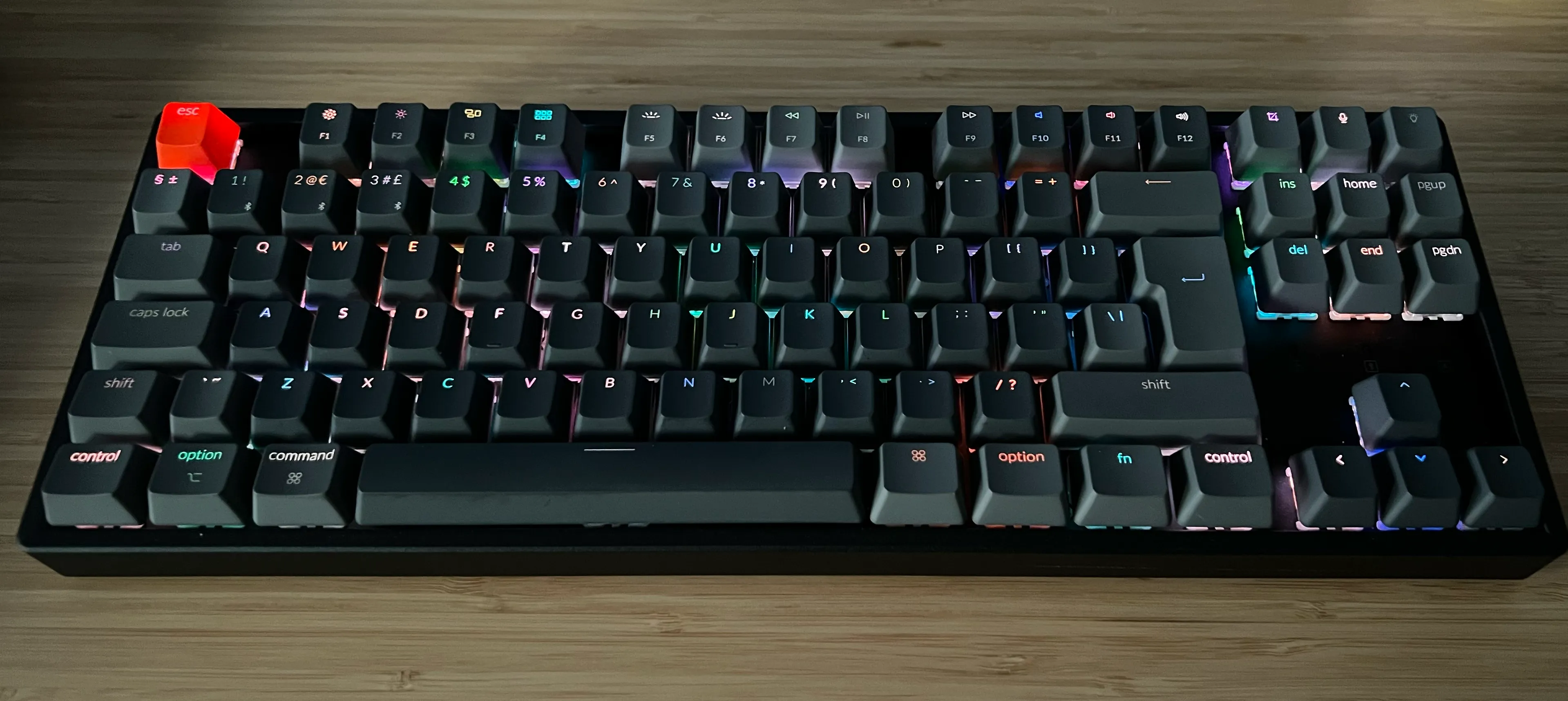 A tenkeyless wireless keyboard with dark grey keycaps, except for the escape key which is orange. The board has RGB backlighting, which is glinting different colours under each key.