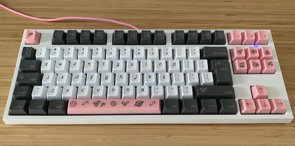 A white mechanical keyboard, with white, pink, and dark grey keycaps. The font on the keycaps is curly and italic. A bunch of the keys have space themed icons.