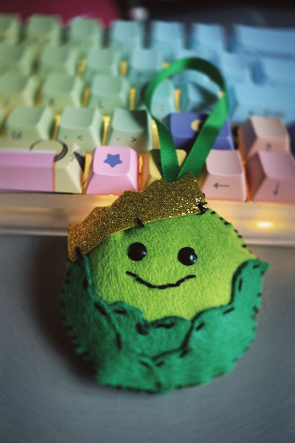 Felt tree ornament of a little green brussel sprout, with a smile and a golden crown.