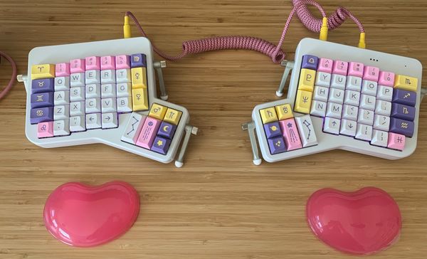 A white mechanical keyboard, split in two, with white, pink, yellow and purple keycaps in an astrological theme.