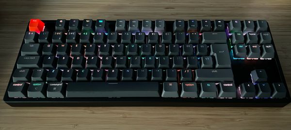 A tenkeyless wireless keyboard with dark grey keycaps, except for the escape key which is orange. The board has RGB backlighting, which is glinting different colours under each key.  